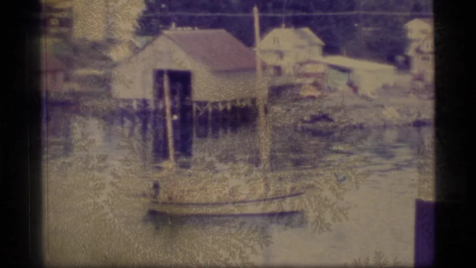 1977 Alaska harbor frame with waterfront buildings, pilings, a small boat, reflective water, and nearby shoreline structures.