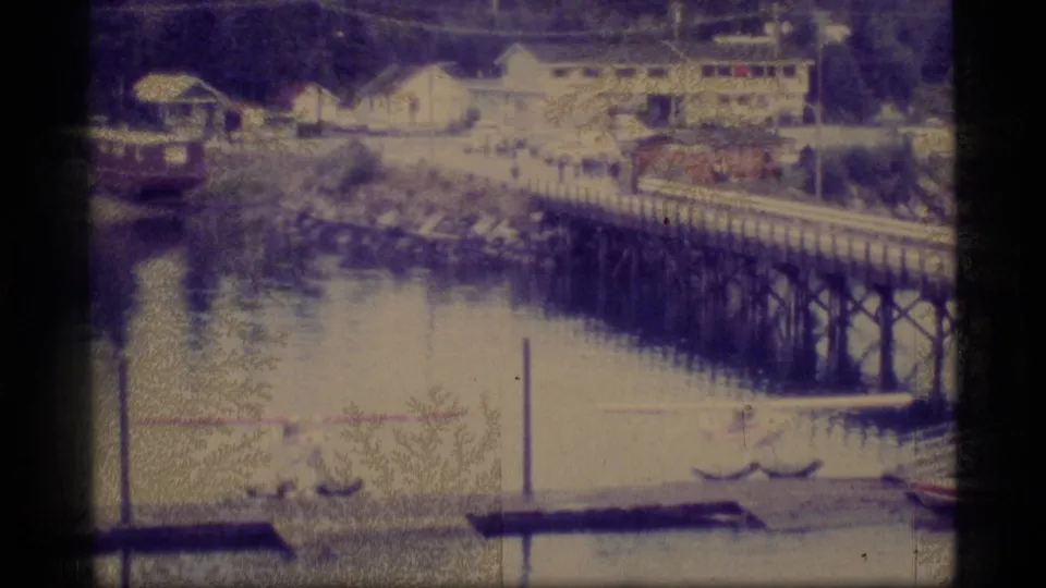 1977 Alaska home-movie frame showing a calm waterfront, dock or trestle, shoreline buildings, a small vessel, and beached floatplanes.
