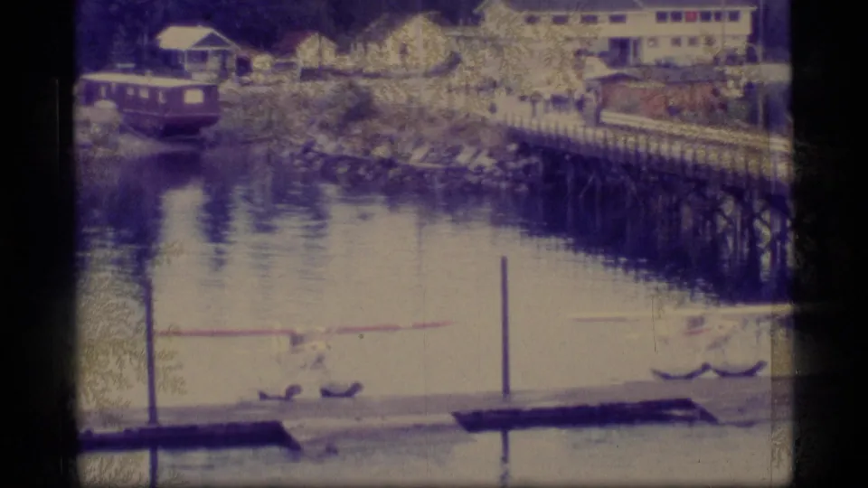 1977 Alaska waterfront view with low buildings, a long timber dock or trestle, calm water, a small vessel, and beached floatplanes.