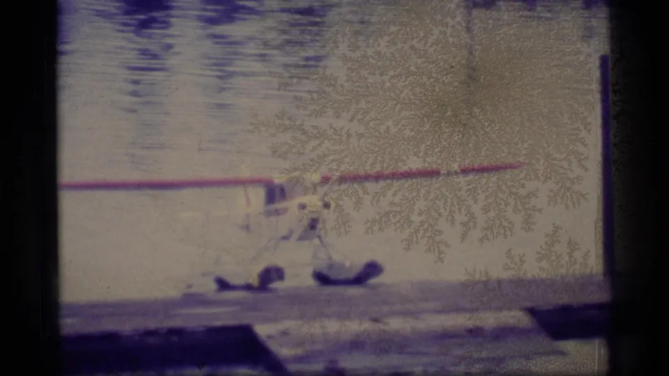 1977 Alaska home-movie frame closer to the shore, showing a red-and-white floatplane wing, aircraft floats, still water, and a dock edge.