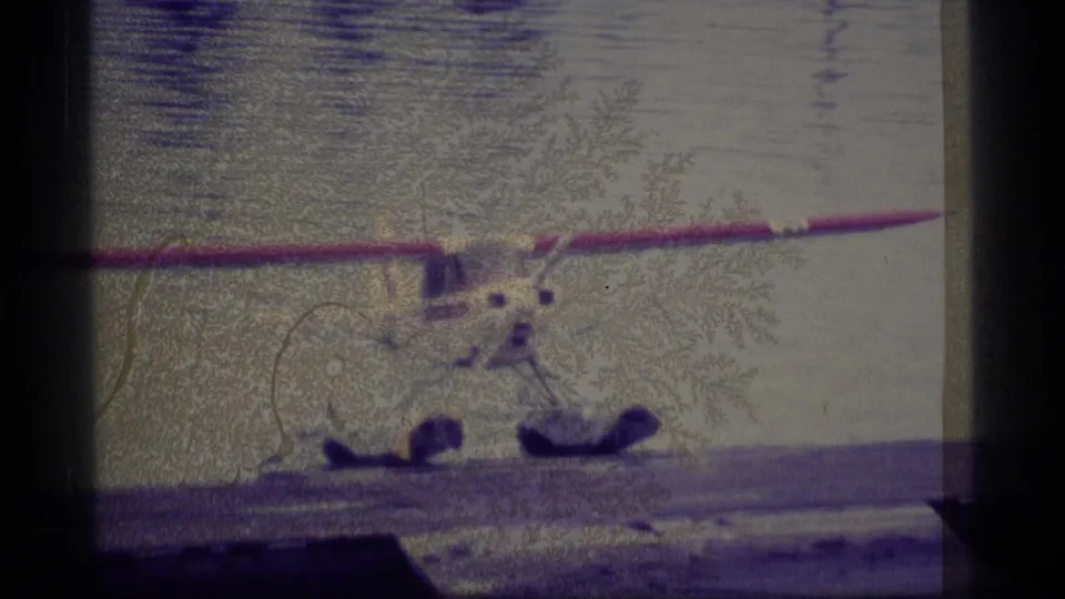 1977 Alaska frame showing beached floatplanes at the water's edge, with a red wing, floats, dock structure, and shoreline vegetation.