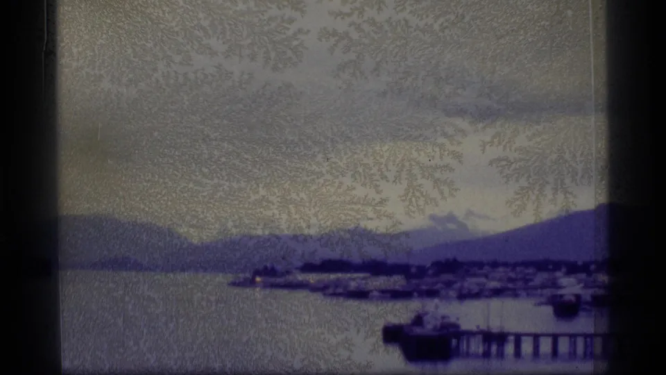 1977 Alaska home-movie frame looking across water toward a harbor town, pier, boats, and layered mountains under soft exposure.