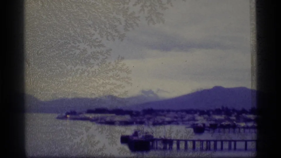Soft 8mm Alaska harbor frame with a pier and boat in front, a distant waterfront settlement, and mountains beyond the water.
