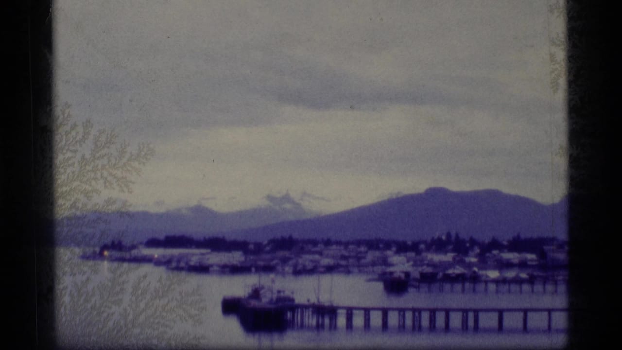 1977 Alaska 8mm frame showing a waterfront pier, small harbor structures, calm bay water, and mountains under a cloudy sky.