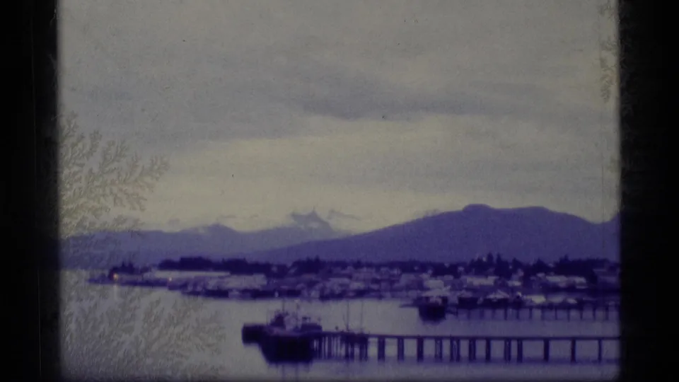1977 Alaska 8mm frame showing a waterfront pier, small harbor structures, calm bay water, and mountains under a cloudy sky.