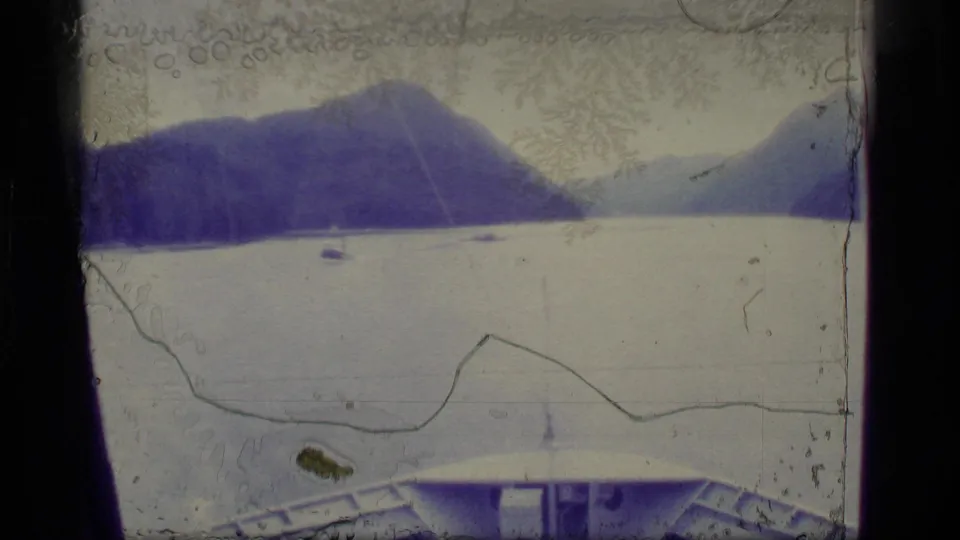 1977 Alaska 8mm frame looking across calm water toward mountain slopes, with small boats and a vessel deck visible in the foreground.