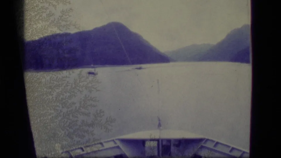 1977 Alaska home-movie frame from a vessel or waterfront position, showing calm water, small boats, and mountains in the distance.