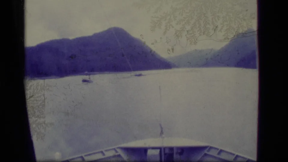 1977 Alaska 8mm frame showing a small boat on a quiet bay, mountain slopes beyond, and part of a vessel deck in the foreground.