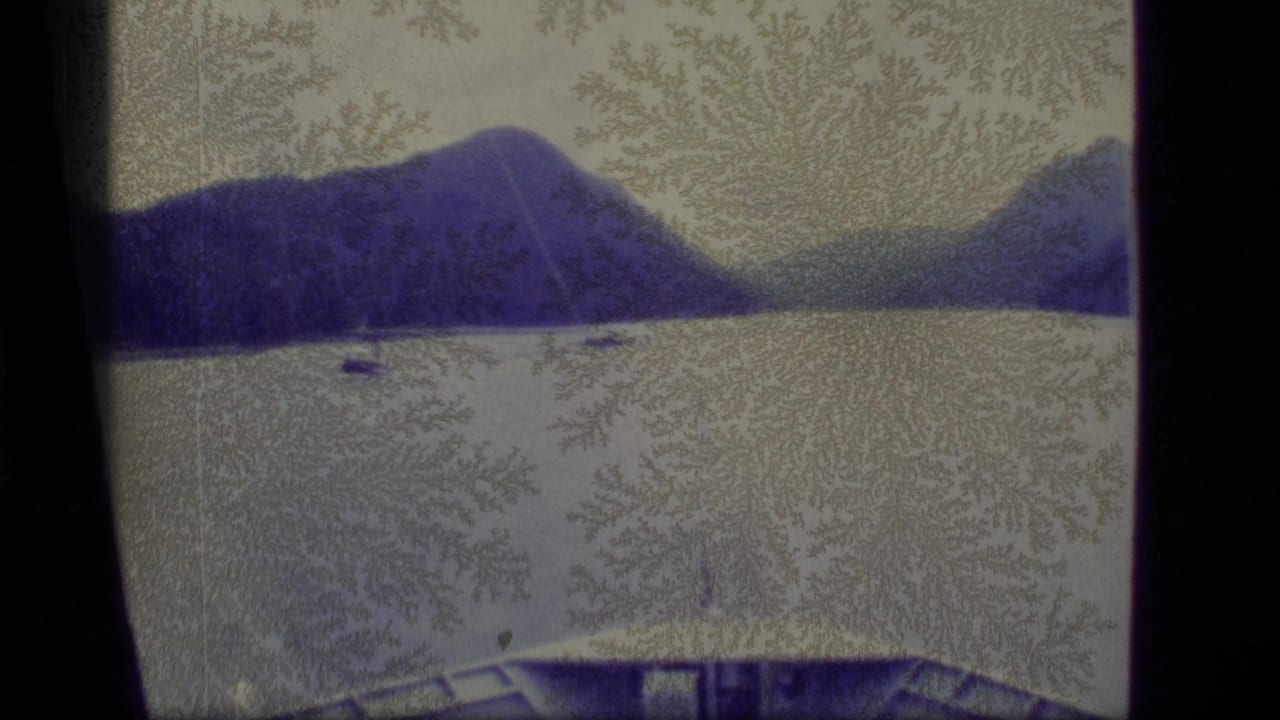 1977 Alaska 8mm frame of a calm lake below blue mountain slopes, with a small distant watercraft and visible surface patterns on the film.