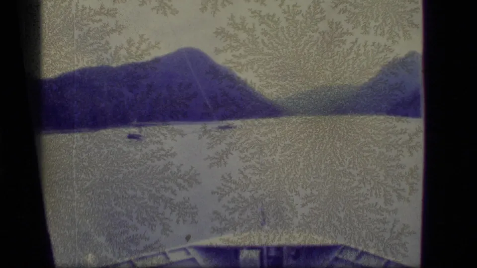 1977 Alaska 8mm frame of a calm lake below blue mountain slopes, with a small distant watercraft and visible surface patterns on the film.