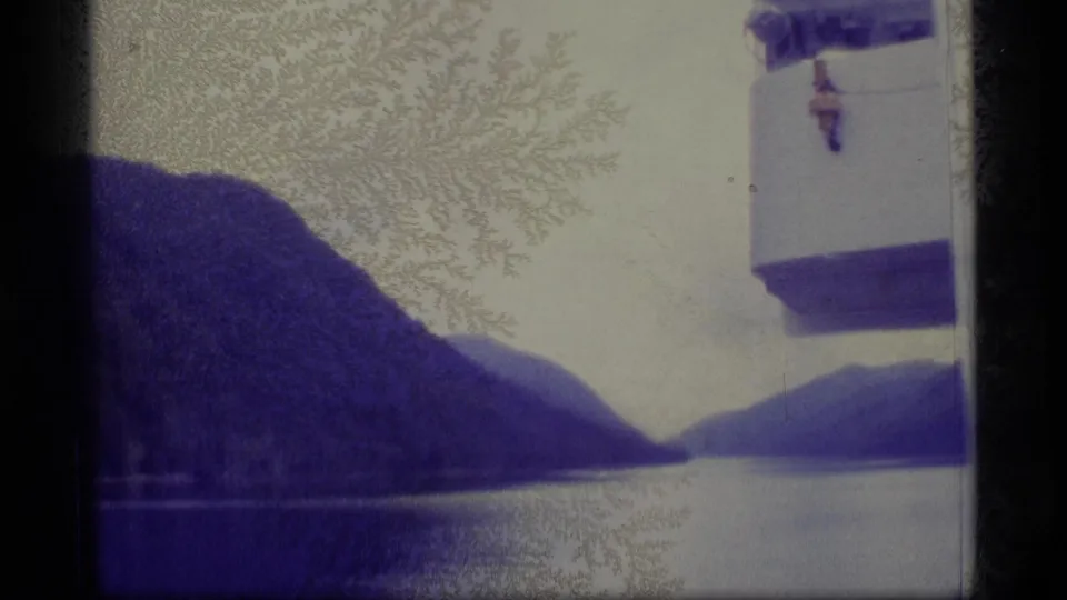 1977 Alaska 8mm frame from a boat trip, with mountain slopes, reflective water, and a white vessel-side structure near the right edge.
