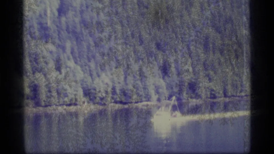 1977 Alaska 8mm frame showing dense forest along a lake, reflected water, and a bright small shoreline structure or boat form.