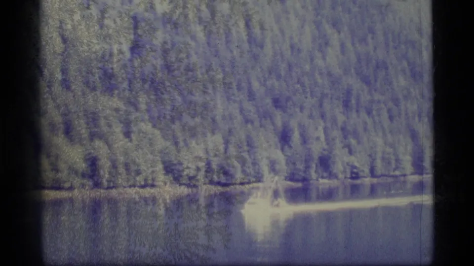 1977 Alaska 8mm frame showing a small boat and wake on calm water beneath a forested shoreline.