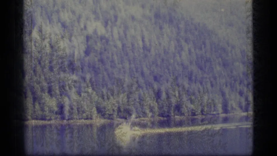 A small boat travels across a calm Alaska waterway in a grainy 1977 8mm frame, with dense trees along the far shore.