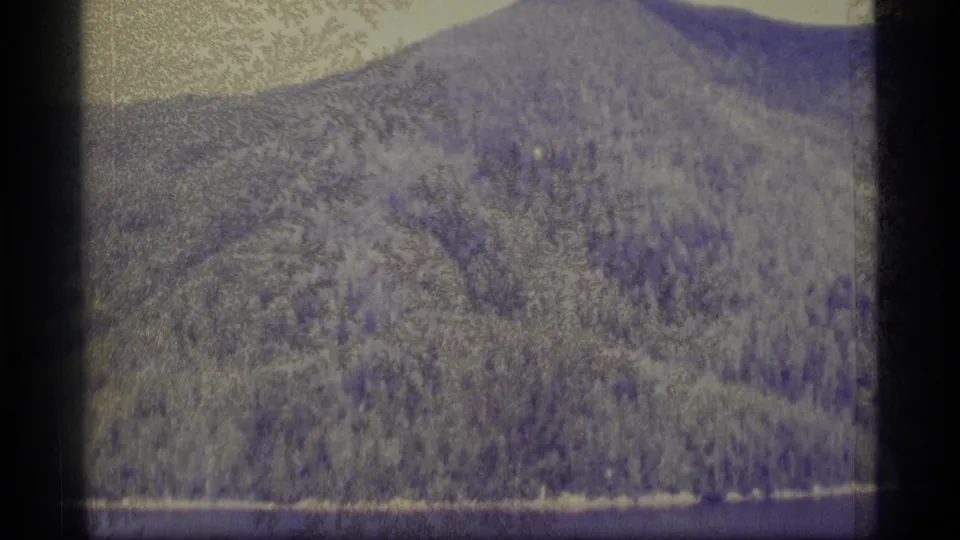 1977 Alaska 8mm frame showing a wooded hillside and calm waterline, with the boat no longer clearly visible.