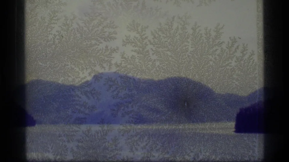1977 Alaska 8mm frame with a mountain ridge beyond calm water, a dark shoreline mass at right, and pronounced aged-film texture.