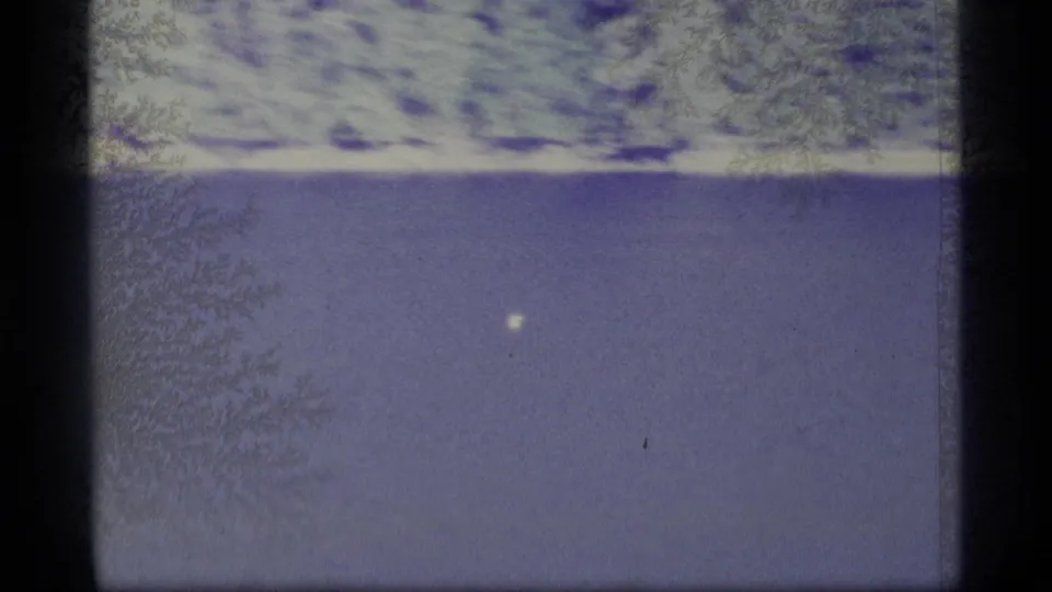 1977 Alaska 8mm frame showing water, a bright distant shoreline band, dark upper texture, and a small white speck that may be a bird.