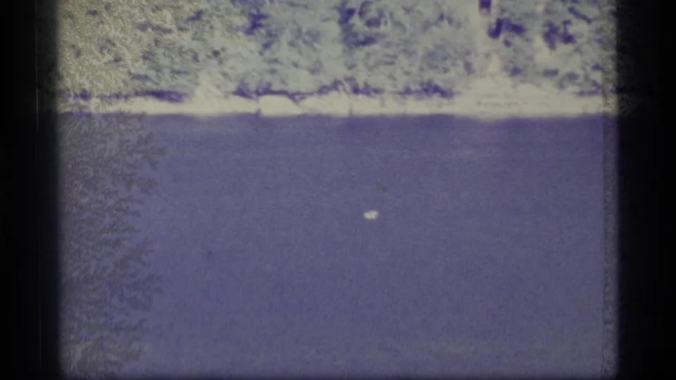 1977 Alaska 8mm frame with blue-toned water, a small white speck near the surface, distant pale shoreline, and visible film wear.