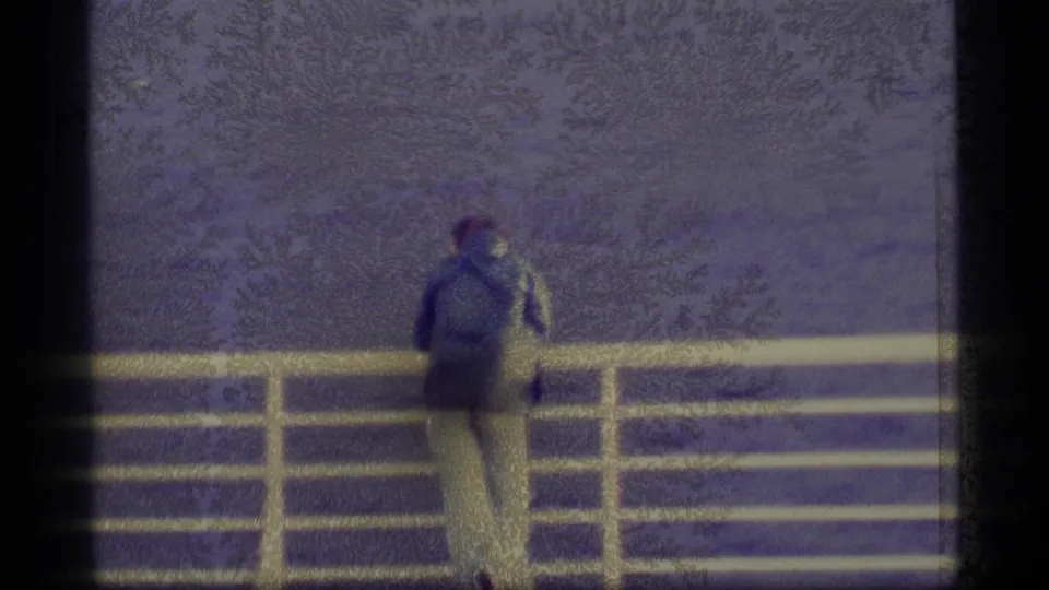 Grainy 1977 Alaska home-movie frame with a distant person at a pier railing, water behind them, and soft tree shapes in the background.