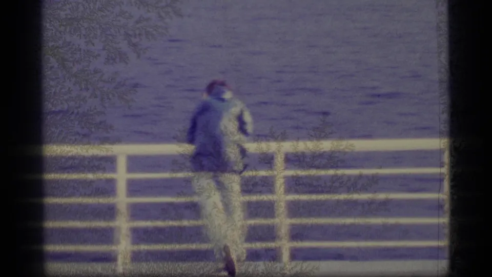 Faded 8mm frame from 1977 Alaska showing a person at the railing beside open water, seen from behind with visible gate edges.