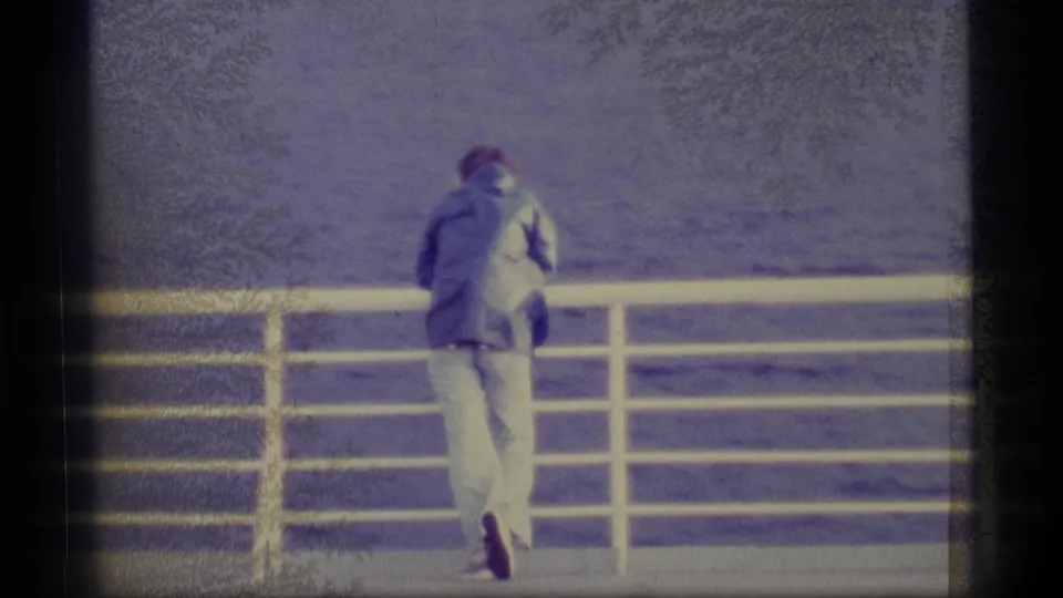 1977 Alaska 8mm frame showing a man in a jacket and light pants at a ship railing, looking over open water.