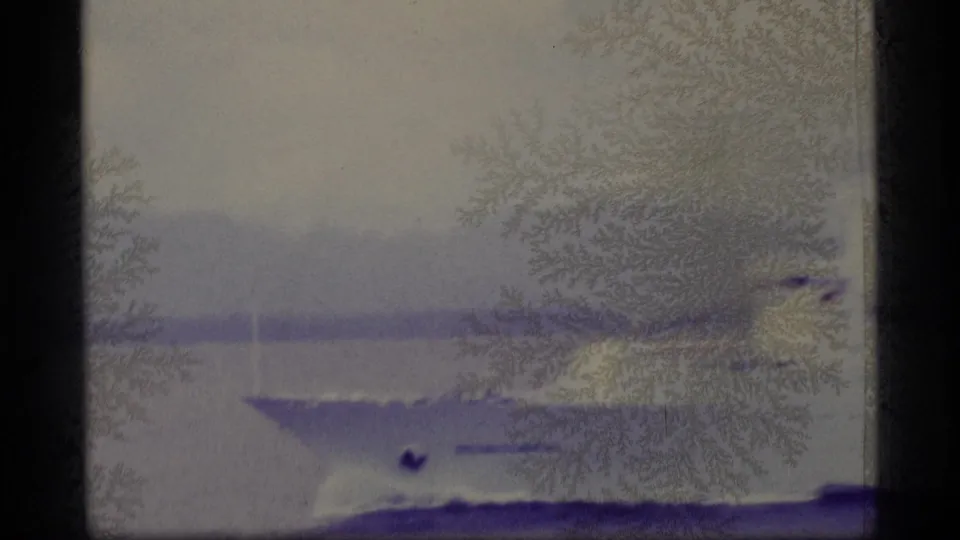 1977 Alaska home-movie frame showing a white passenger ship on calm coastal water under heavy purple staining and emulsion damage.