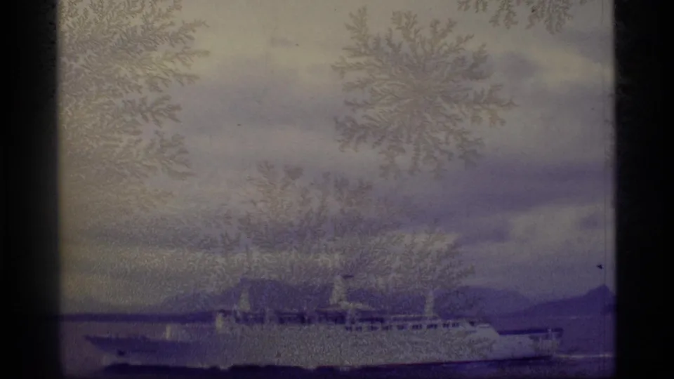 1977 Alaska home-movie frame with a distant passenger ship crossing coastal water, partially veiled by frost-like film patterns.
