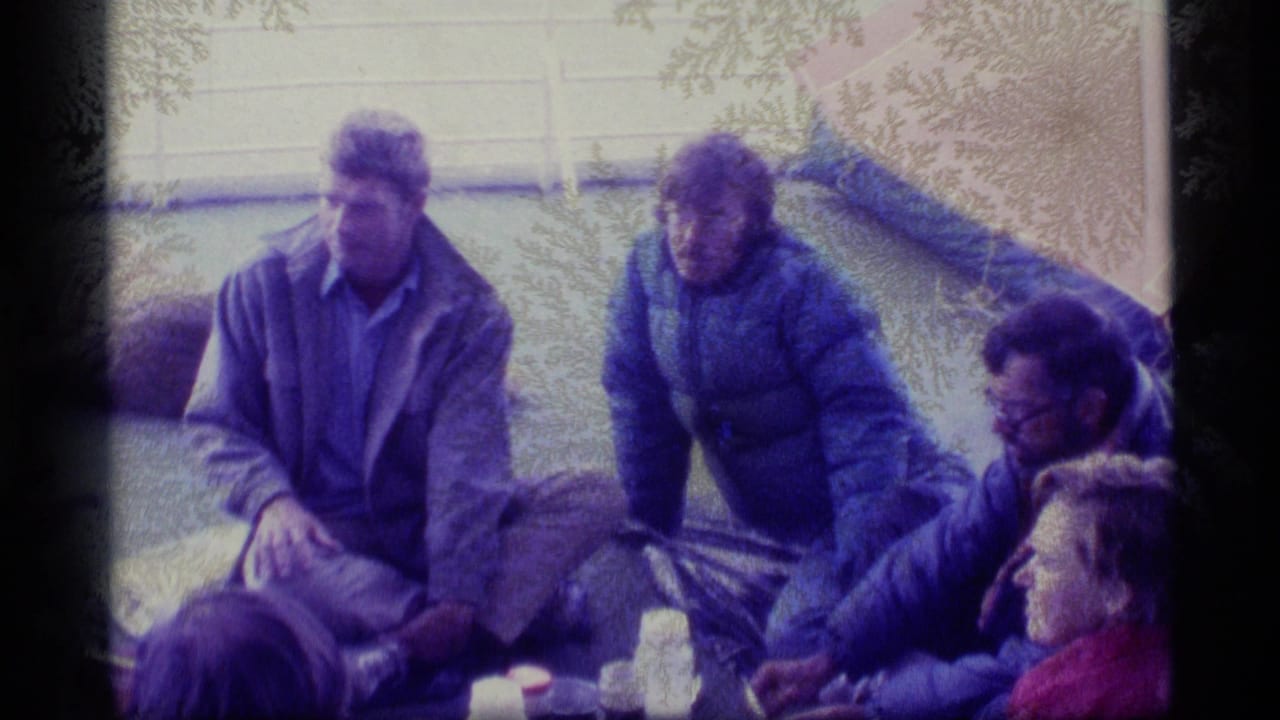 1977 Alaska home-movie frame of adults in cold-weather jackets seated on a deck near bags and blankets.
