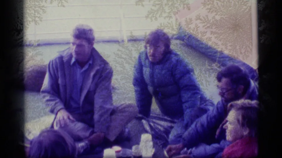 1977 Alaska home-movie frame of adults in cold-weather jackets seated on a deck near bags and blankets.
