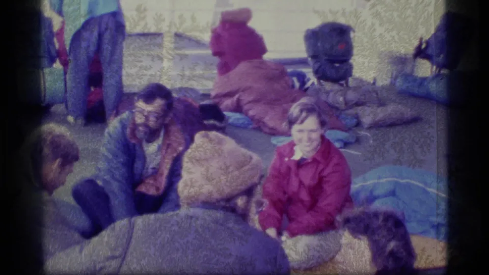 1977 Alaska home-movie frame showing travelers resting on a deck with outdoor gear nearby.