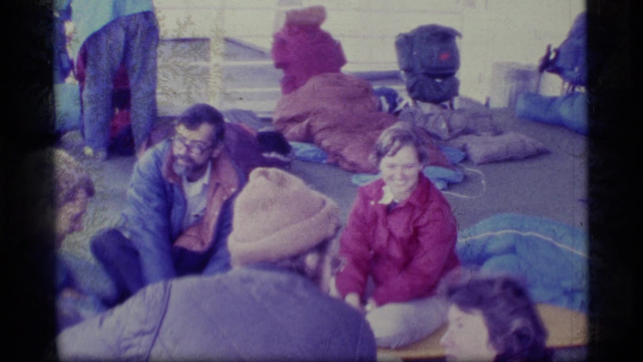 Adults in coats sit in a circle on a boat deck with bags and bedding nearby.