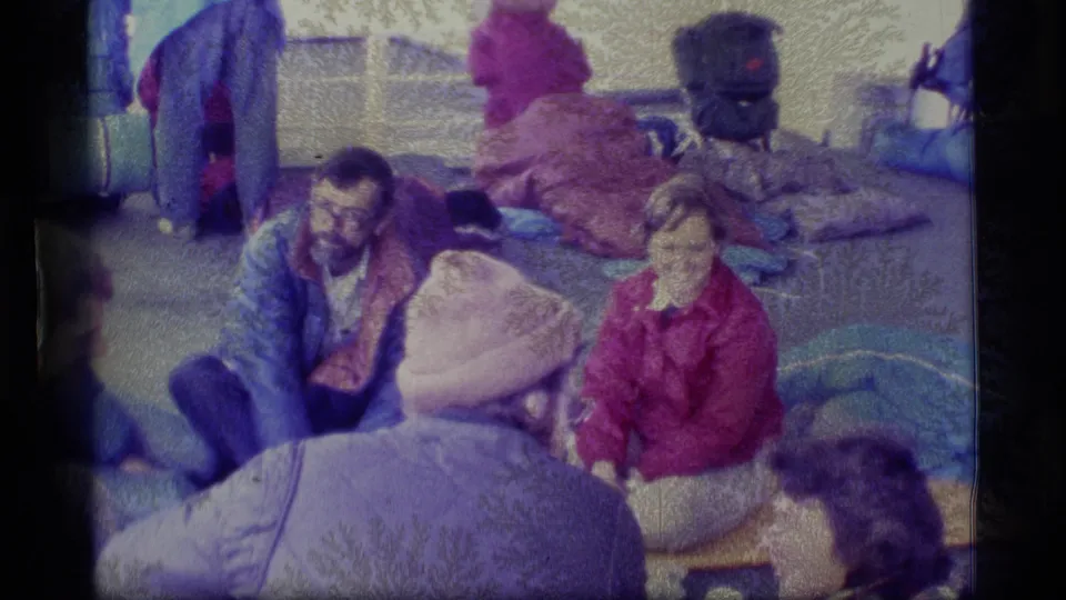 Bundled adults appear to converse on an Alaska boat deck, surrounded by soft luggage and sleeping bags in faded 8mm color.