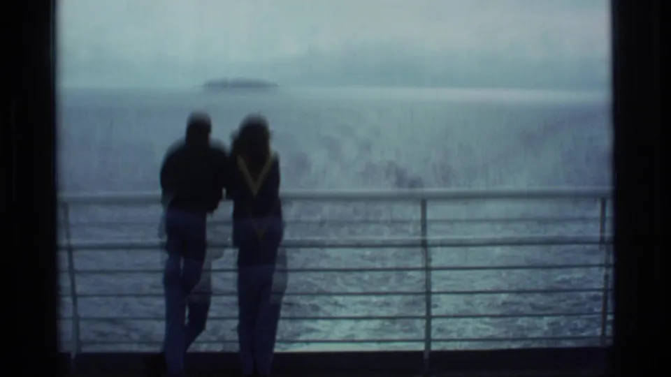 Two silhouetted travelers stand at a boat railing, looking across Alaska coastal water toward a low island under cloudy sky.