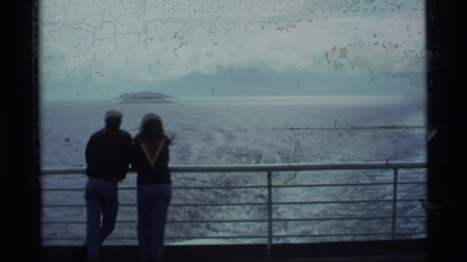 A pair of travelers stands together at a railing as the 8mm frame shows Alaska water, a distant island, and overcast light.