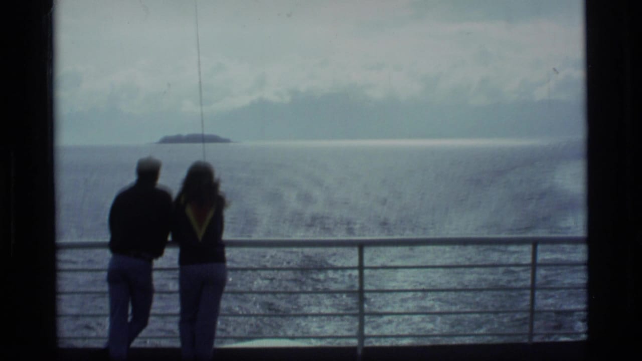 In 1977 Alaska cruise footage, two adults appear in silhouette at a deck rail with open water, horizon, and a distant island beyond.
