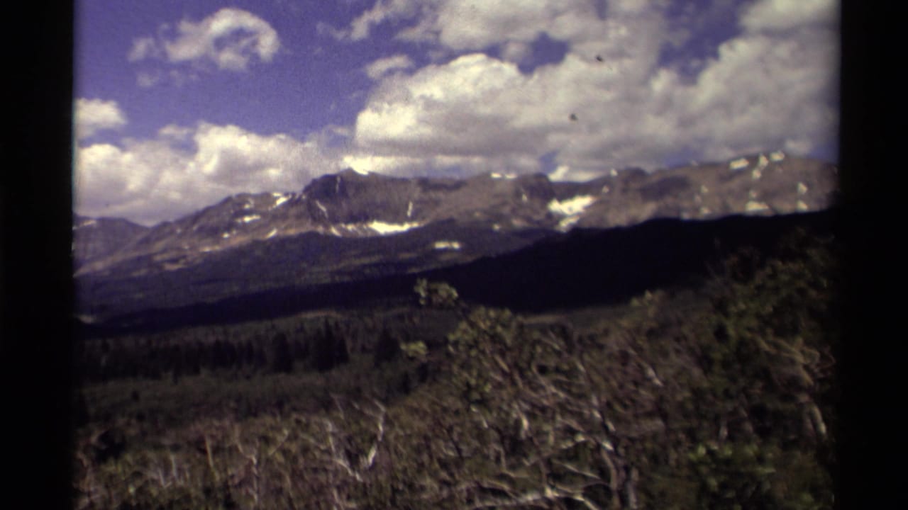 1979 8mm Canada home-movie frame showing rugged mountains, forest, brush, blue sky, and scattered white clouds.