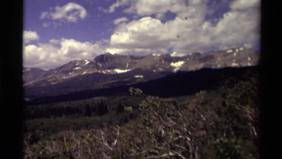 1979 8mm Canada home-movie frame showing rugged mountains, forest, brush, blue sky, and scattered white clouds.