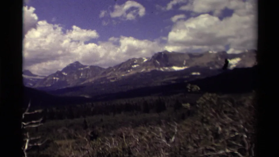 1979 8mm Canada frame with mountain ridges, a forested valley, brush, blue sky, clouds, and dark scan borders.