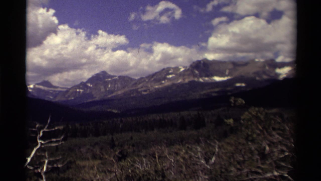 Snow-dusted mountain range below a blue sky with clouds and dark forest in the foreground.