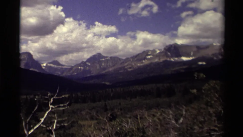 1979 Canadian 8mm frame with a broad mountain ridge, scattered snow patches, dense forest, bright clouds, and a dark scan edge.