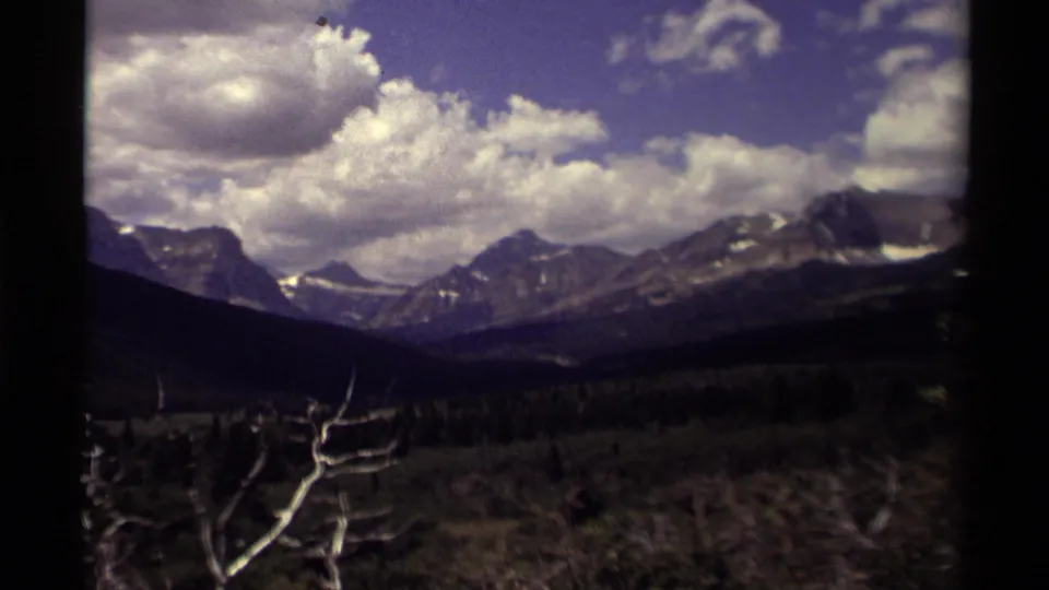 1979 Canadian 8mm frame showing distant peaks, a large cloud bank, dark forested slopes, and pale bare branches in the lower frame.