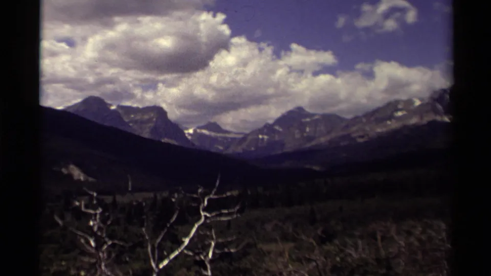1979 Canadian 8mm frame with snowy peaks, layered dark forest, bright white clouds, blue sky, and foreground branches.