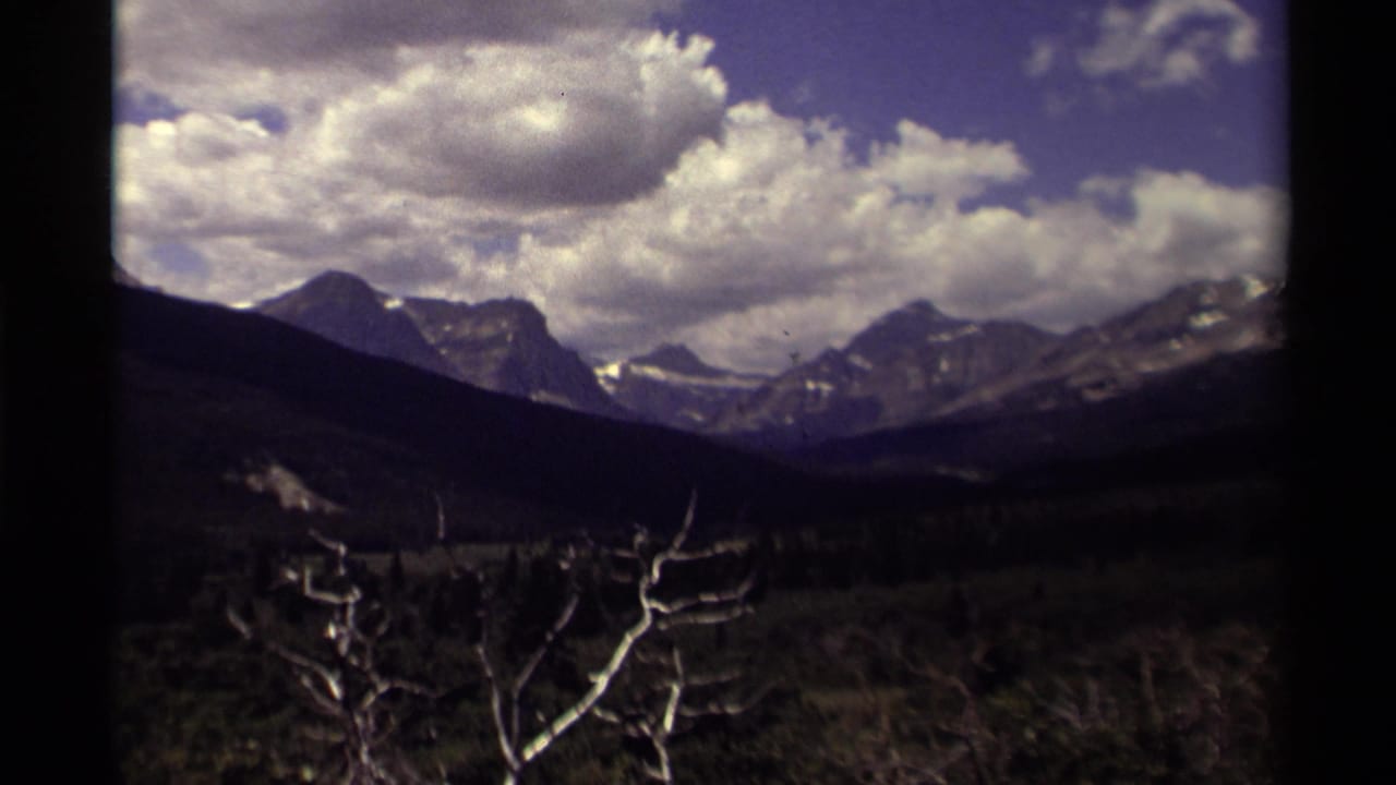 1979 Canada 8mm frame showing distant rugged mountains beneath large clouds, with a dark evergreen valley and bare branch in the foreground.