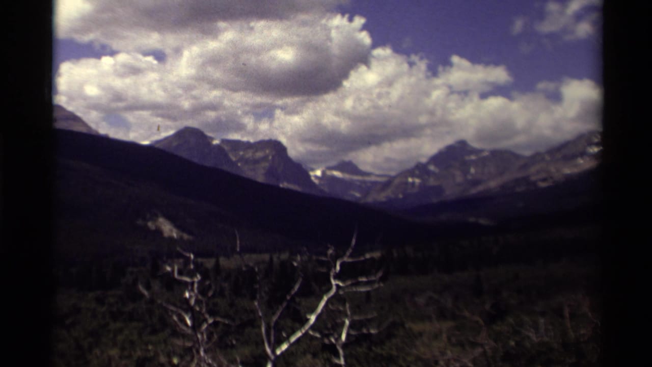 1979 Canada home-movie frame of broad mountain peaks under billowing clouds, with shadowed valley slopes and pale bare branches in front.