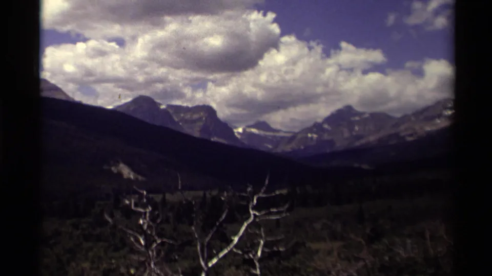 1979 Canada home-movie frame of broad mountain peaks under billowing clouds, with shadowed valley slopes and pale bare branches in front.