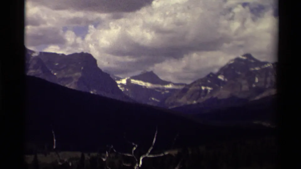 1979 Canada 8mm frame with rugged peaks, small snowfields, dense clouds, a dark valley foreground, and bare branches near the lower edge.