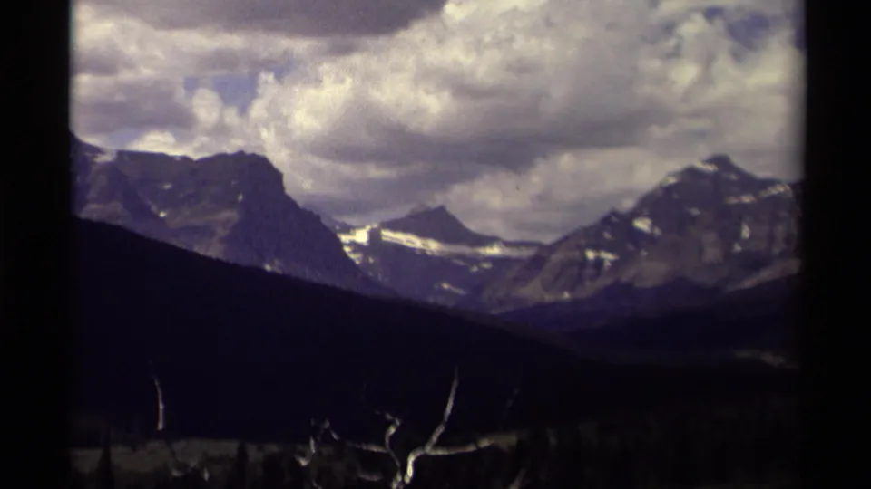 1979 Canada home-movie frame showing heavy clouds above rugged mountain peaks, a shadowed valley below, and bare branch tips near the foreground.