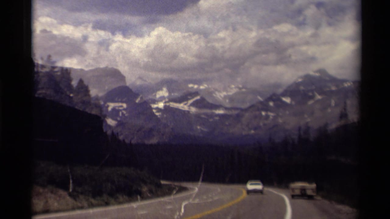 1979 Canadian Rockies road view with a curving highway, evergreen forest, distant snow-streaked mountains, and two vehicles ahead.
