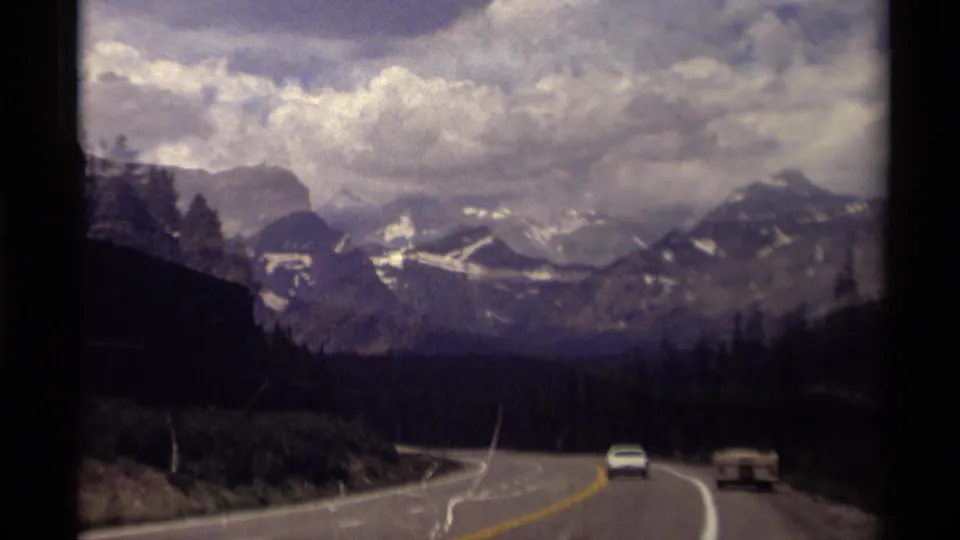 1979 Canadian Rockies road view with a curving highway, evergreen forest, distant snow-streaked mountains, and two vehicles ahead.
