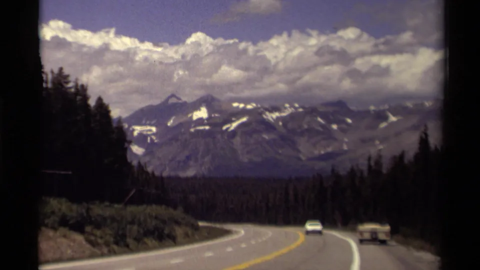 1979 Canadian road-trip frame showing a forest-lined highway, white car, small trailer, and snow-streaked mountains under bright clouds.